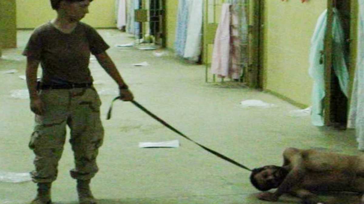 Disturbing image of woman in military fatigues with a naked man on the floor, leashed around his neck.